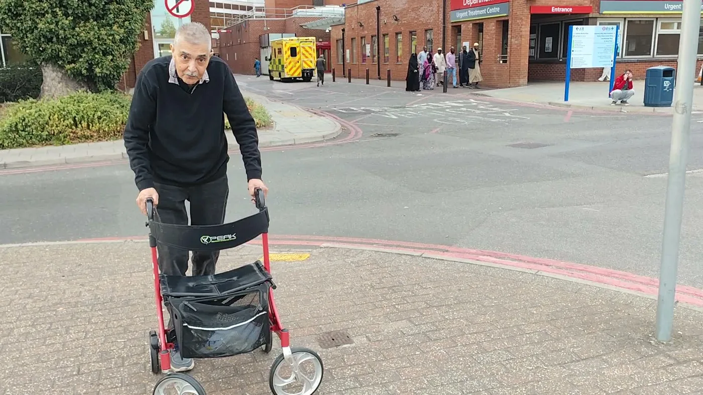A man with a red wheeled walking frame on the pavement opposite a hospital entrance